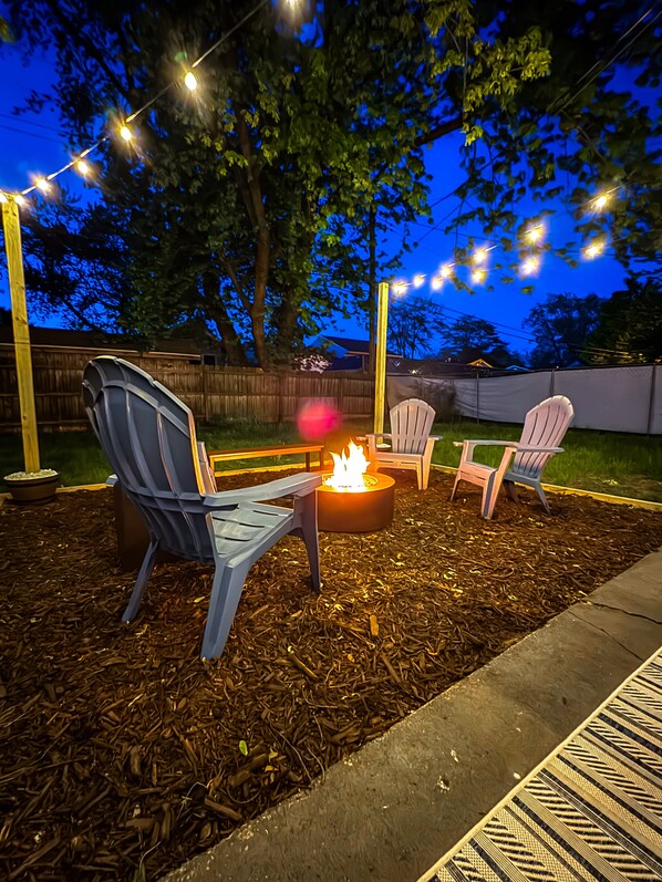 Un espacio al aire libre con sillas Adirondack y una chimenea, rodeado de mantillo e iluminación con luces.