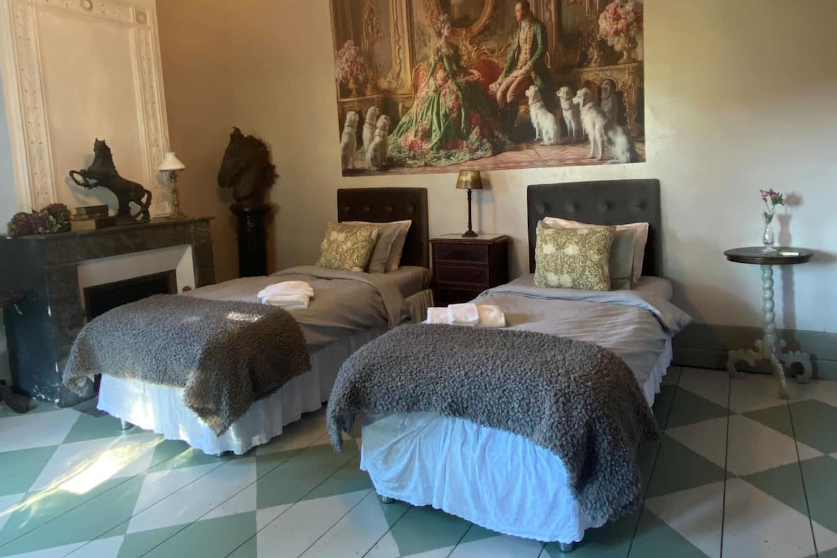 A room with two beds, a chandelier, a large painting, and a checkered floor.