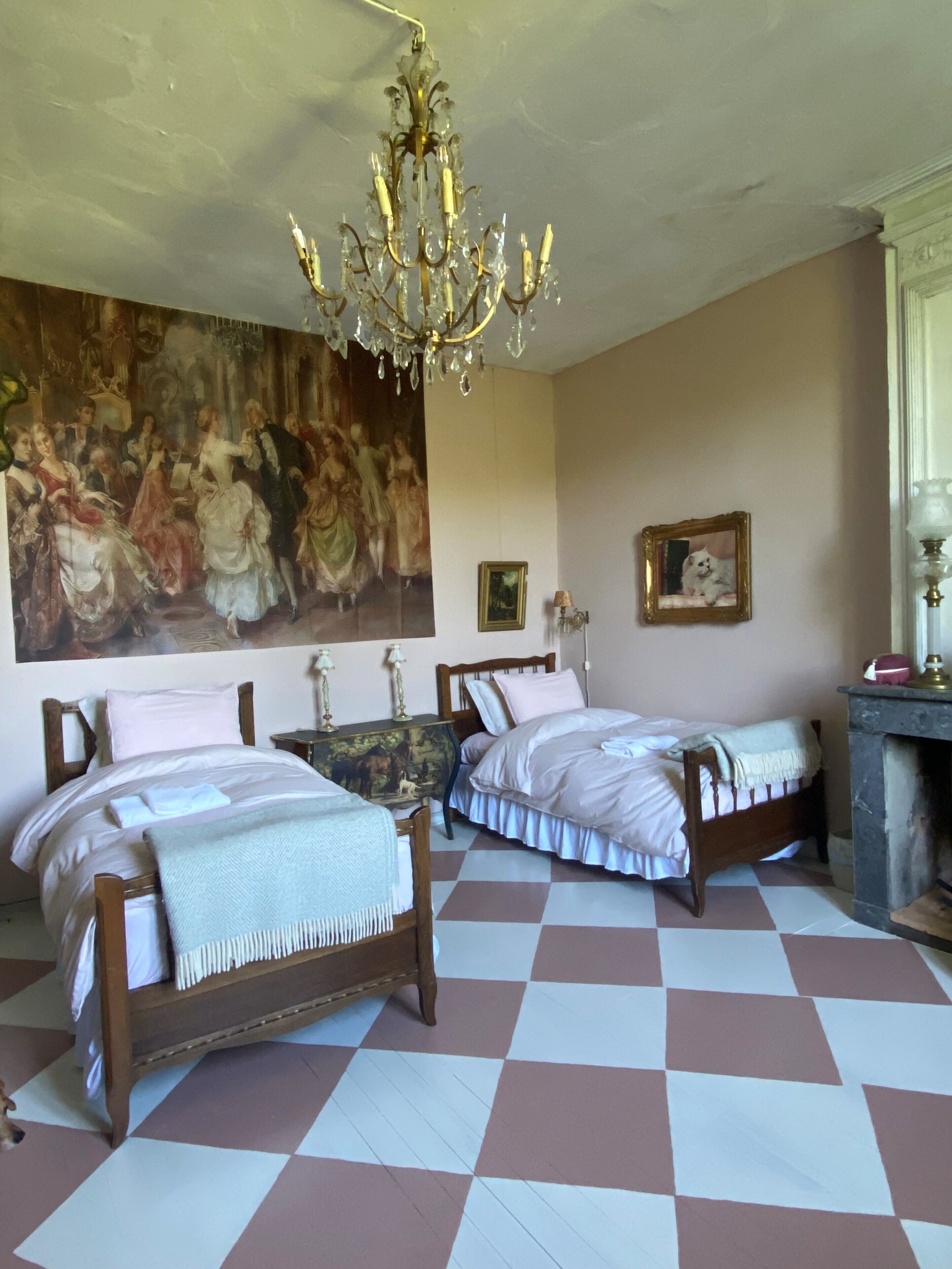 A bedroom with two wooden beds, a large painting on the wall, a chandelier, and a fireplace.