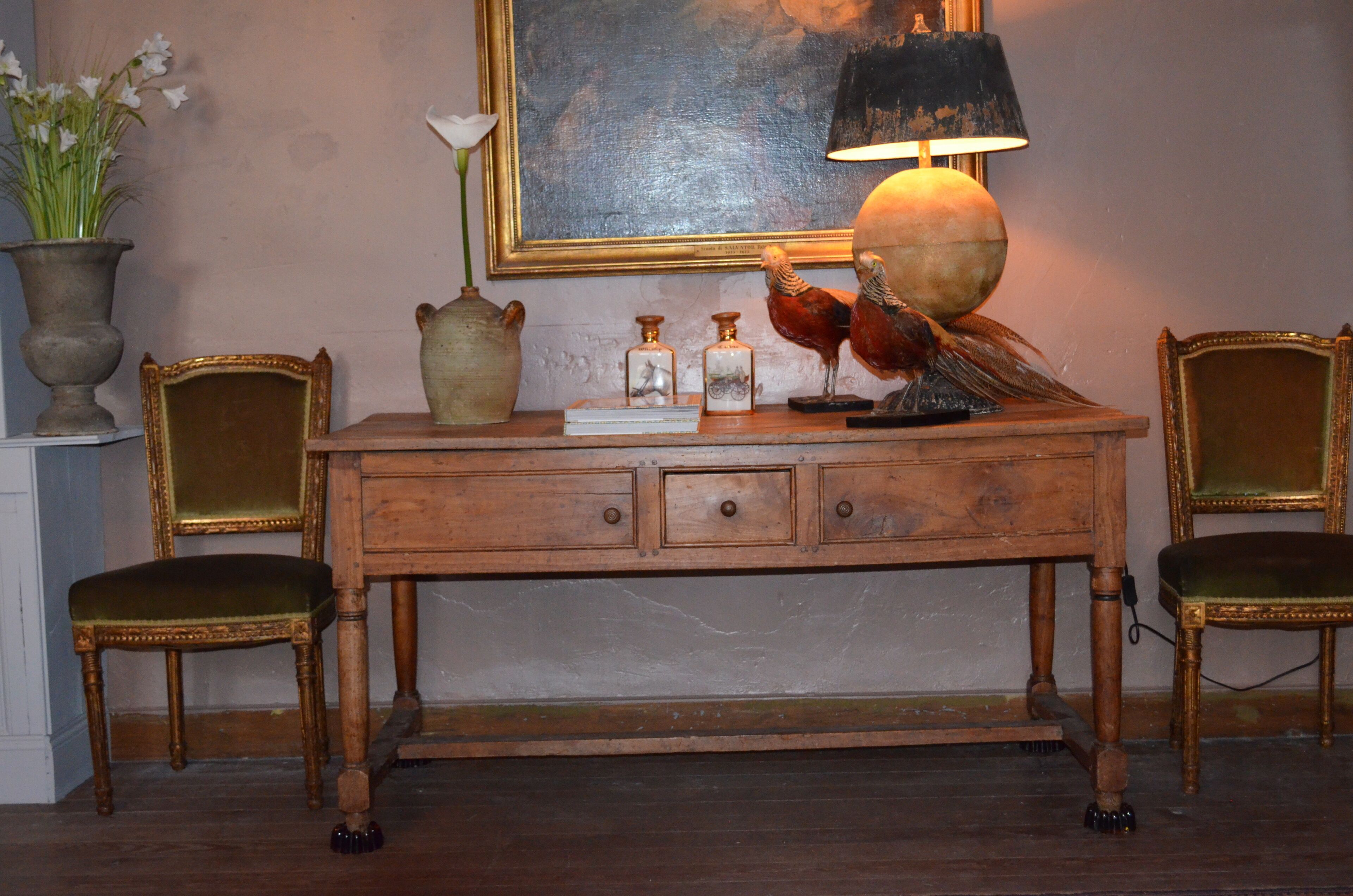 Een houten bureau met lades, een vaas met bloemen, een lamp en twee stoelen.