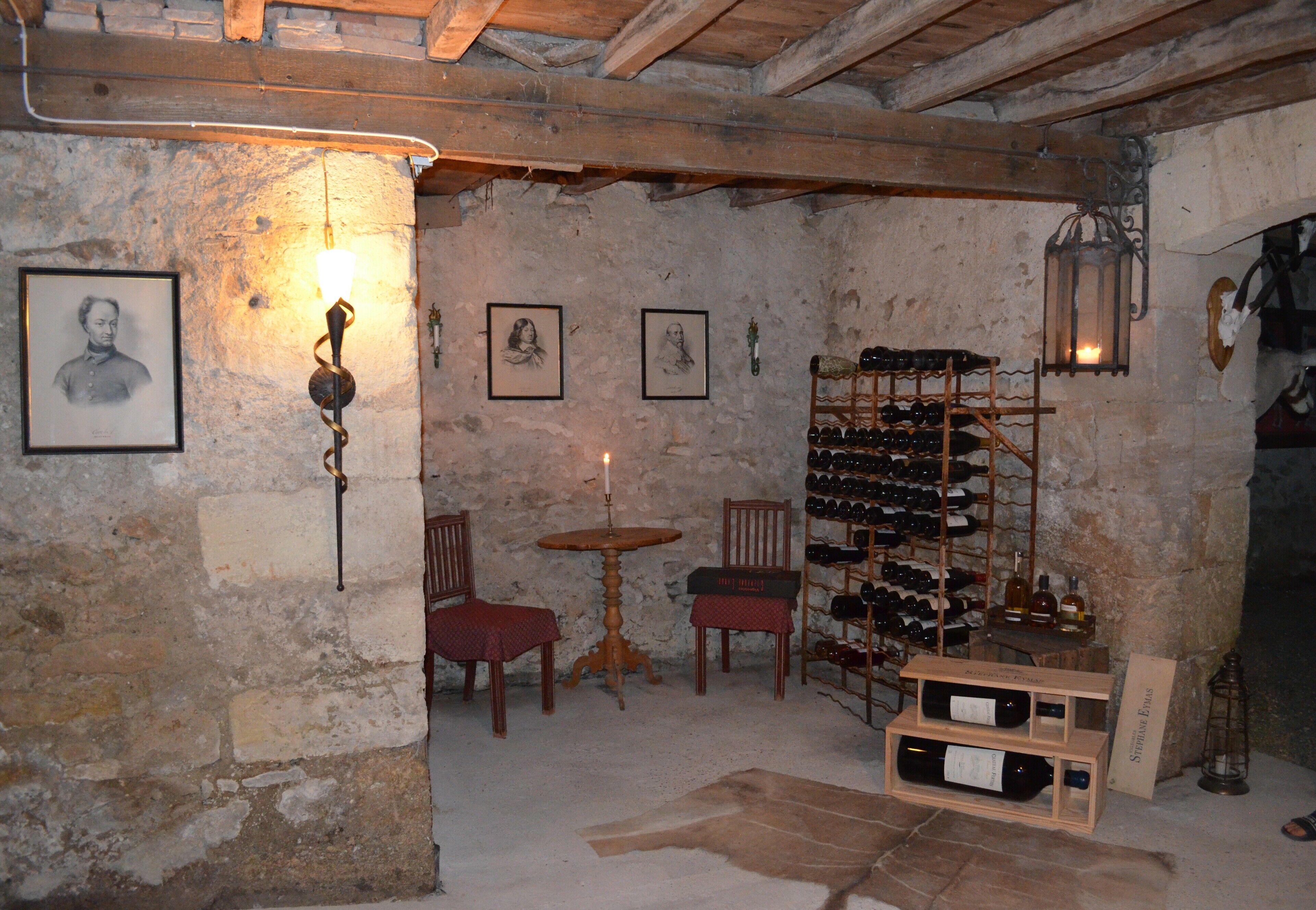 A rustic cellar with stone walls, wooden furniture, a wine rack, and framed portraits.