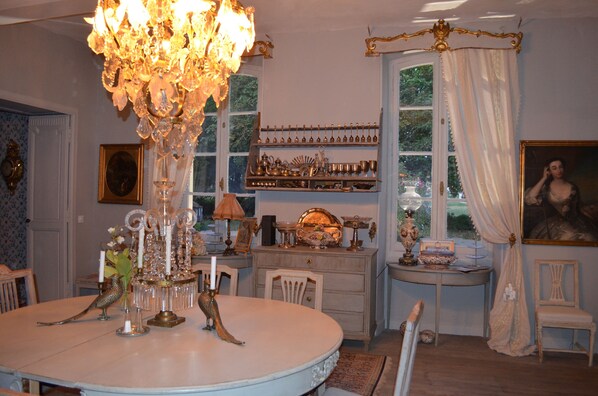 Un comedor con candelabro, una mesa redonda, sillas, una estantería con adornos y una ventana con cortinas.