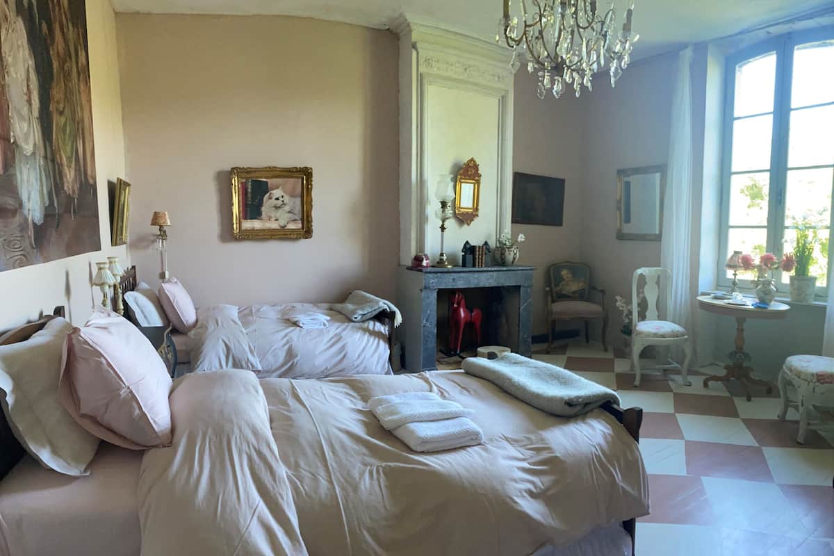 A bedroom with two beds, a fireplace, a mirror, a chandelier, and a window with a view.