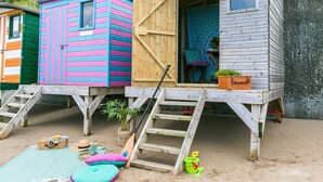 A row of colorful wooden cabins with a sandy ground, a ladder, and beach accessories.
