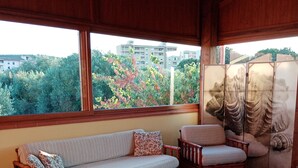 A room with a sofa, two armchairs, and two small tables. There is a view of trees and buildings through large windows.