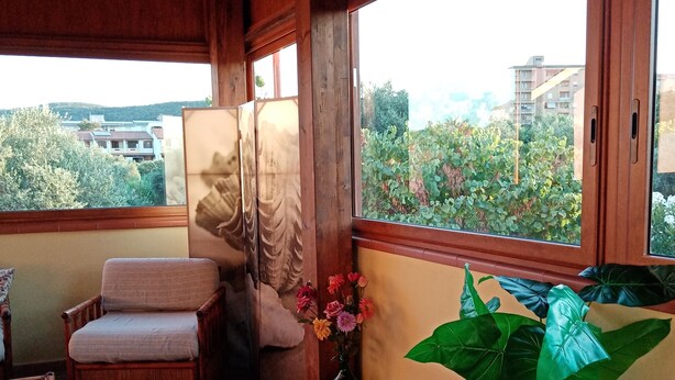 A balcony with a wooden ceiling, a table with a floral tablecloth, a chair, a potted plant, and a vase with flowers.