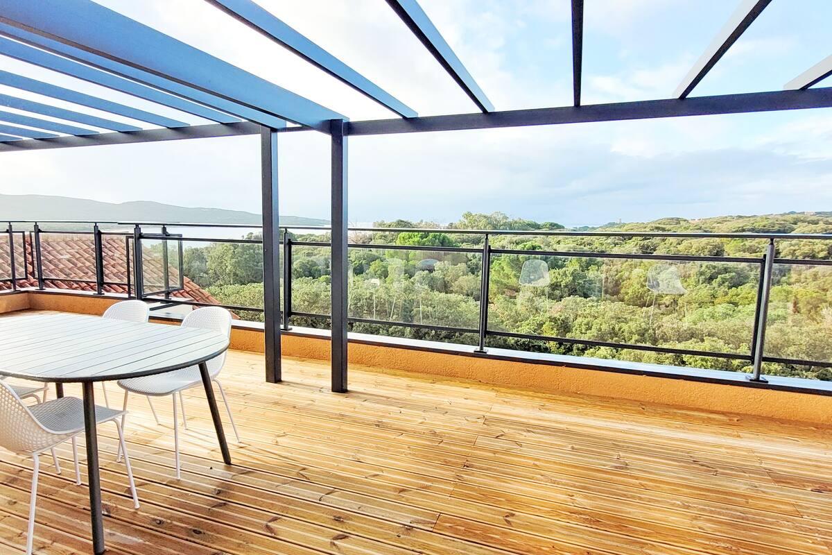 Una terraza de madera con una mesa y sillas, barandilla de cristal y vista a un paisaje verde.