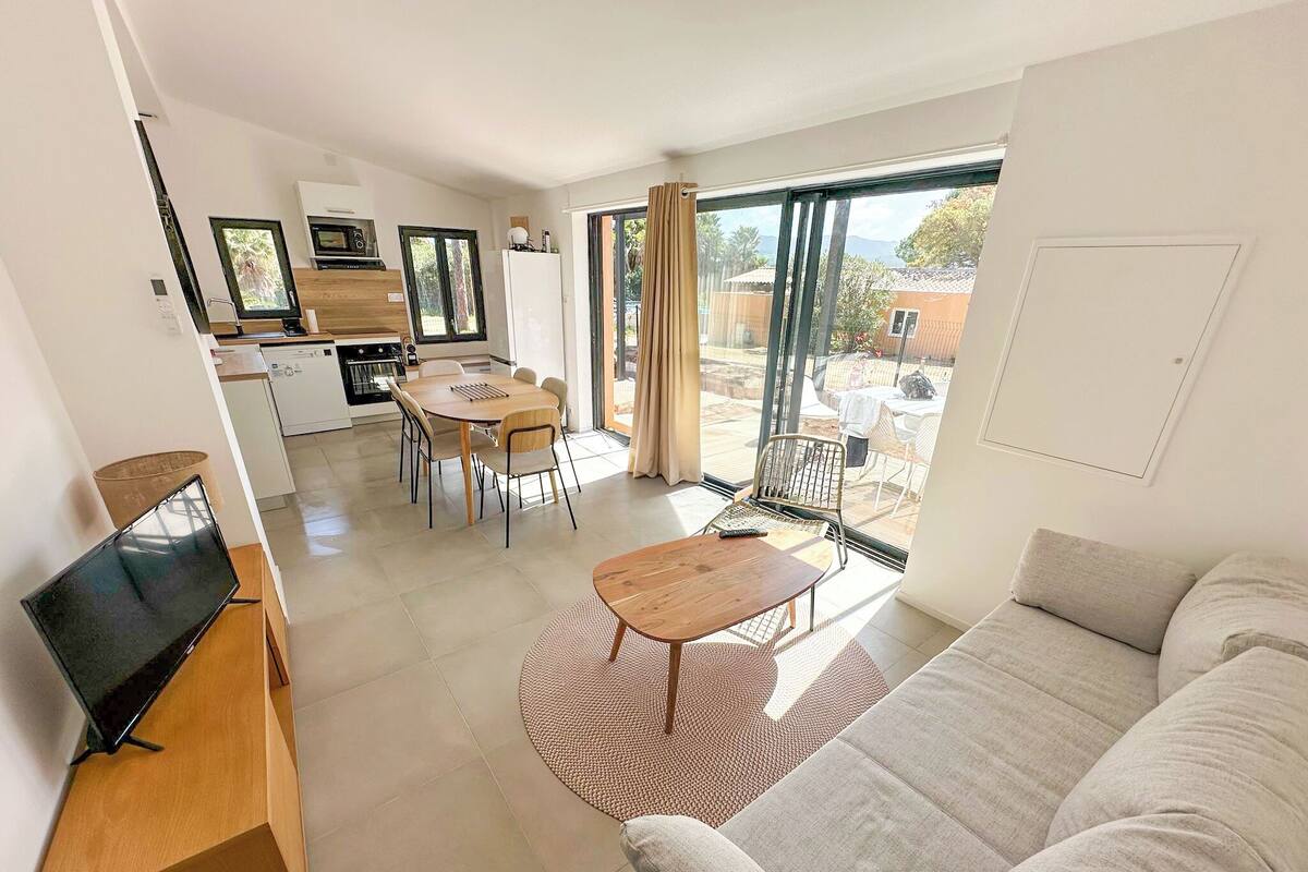 Una sala de estar moderna con un sofá, un mueble para la TV, un comedor con una mesa y sillas, y una cocina al fondo.