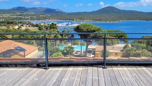 Une terrasse en bois dotée d’une balustrade en verre, offrant une vue sur une marina et un paysage côtier.