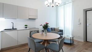 A modern kitchen with white cabinets, a black faucet, and a small potted plant. There's a dining area with a round wooden table, grey chairs, and a vase with flowers. - Italianway Comfort - Cialdini 85 (Milano)