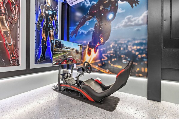 A racing simulator setup with a steering wheel and pedals, a monitor displaying a racing game, and posters of Iron Man on the wall.