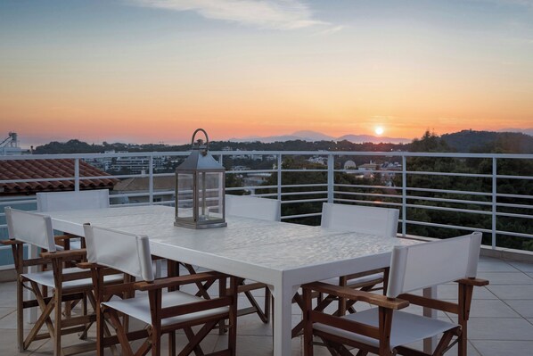 A terrace with a white table and wooden chairs, overlooking a sunset with mountains in the background.