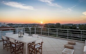 A terrace with a table and chairs, a lantern, and a view of the sunset over a cityscape.