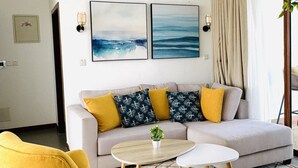 A modern living room with a sofa, yellow armchair, and a coffee table. There are two framed artworks on the wall, a ceiling fan, and a window with curtains. - Serene 2bedroom Beach Apartment, Sultanpalace Kikambala. Northcoast Kilifi Kenya (Kikambala)