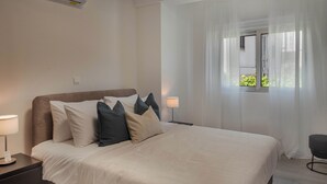 Ein ordentlich bezogenes Bett mit weißer Daunendecke und mehreren Kissen, ein Nachttisch mit Lampe und ein Fenster mit transparenten Vorhängen.