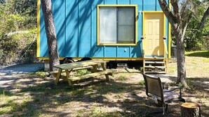 A small, blue and yellow cabin with a window and a door, a picnic table, and a bench in a wooded area. - Tiny House Retro Cabin (Umatilla)