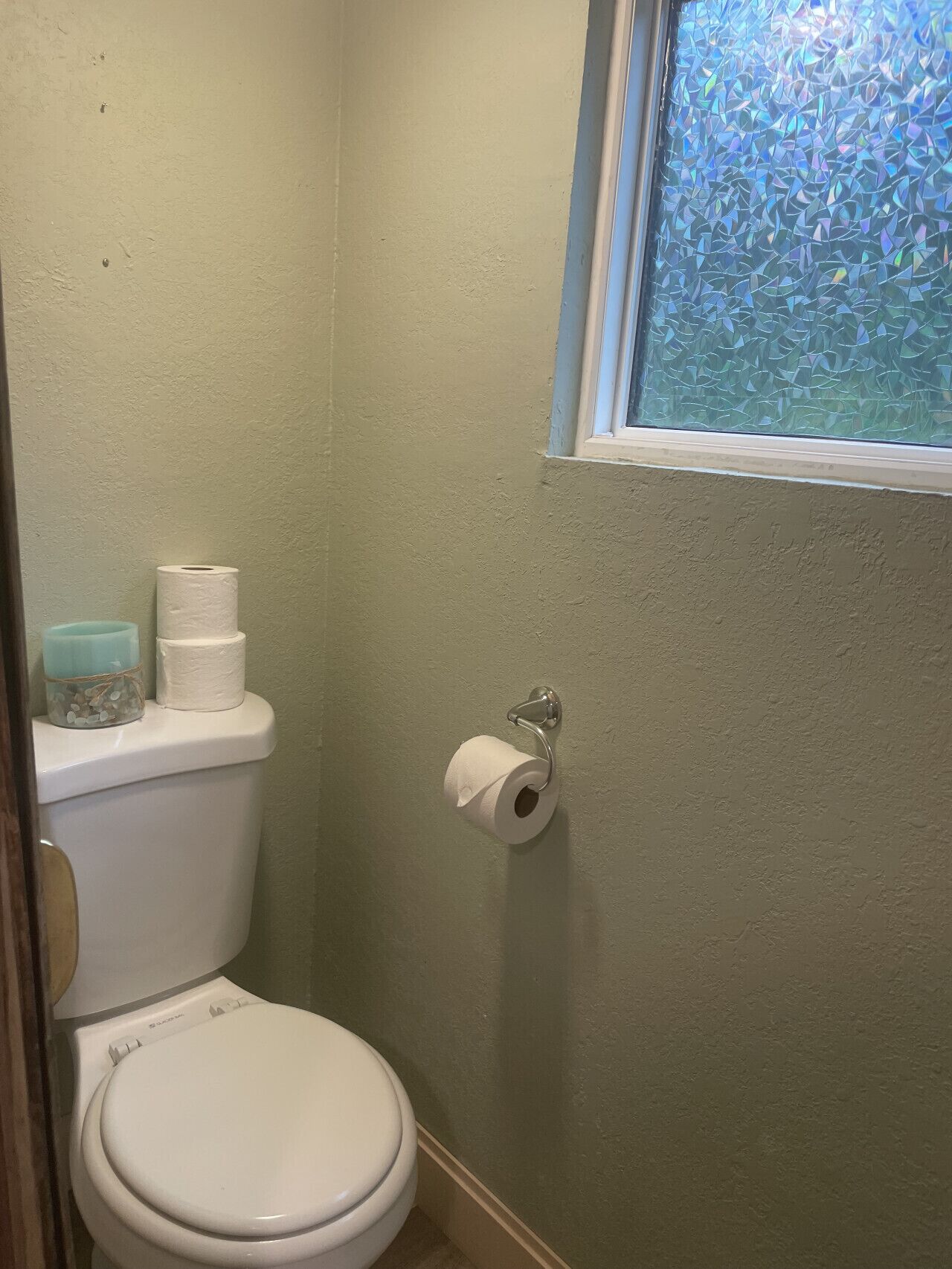 Un baño pequeño con inodoro, dispensador de papel higiénico y ventana con vista a árboles.