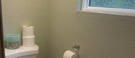 Un baño pequeño con inodoro, dispensador de papel higiénico y ventana con vista a árboles.