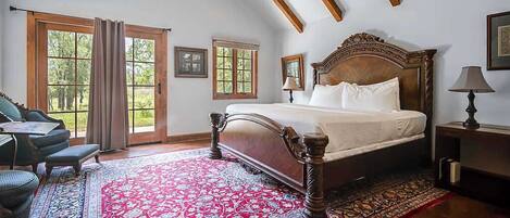 A bedroom with a large bed, a desk, and a chair, with a view of trees through the windows.