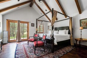 Una cama con dosel, un sillón tapizado rojo y un suelo con alfombra estampada en una habitación con vigas de madera y ventanas amplias.