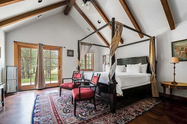 A four-poster bed with a canopy, a red upholstered chair, and a patterned rug in a room with wooden beams and large windows.