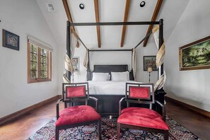 Une chambre à coucher avec un lit à baldaquin, deux fauteuils rouges à revêtement en tissu, un tapis à motifs et des œuvres d’art accrochées aux murs.