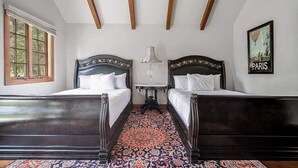 Een kamer met twee houten bedden, een kamerbreed tapijt met dessin, een tafel met een lamp, en een lijst met het woord 'PARIS' op de muur.