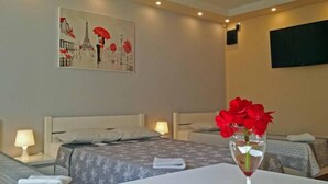 Ett hotellrum med två sängar, en tavla som föreställer en stadsbild, en tv som är monterad på väggen och en vas med röda blommor på bordet.