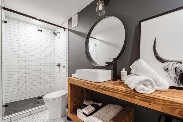 Un baño moderno con inodoro blanco, tocador de madera con espejo y zona de ducha con azulejos blancos.