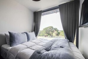 A bedroom with a bed, grey curtains, and a view of greenery outside.
