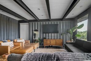 A modern living room with a large TV, leather sofas, a wooden cabinet, and a large window with a view.