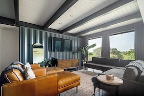 A modern living room with a large TV, leather sofas, a wooden cabinet, and a view of greenery outside.