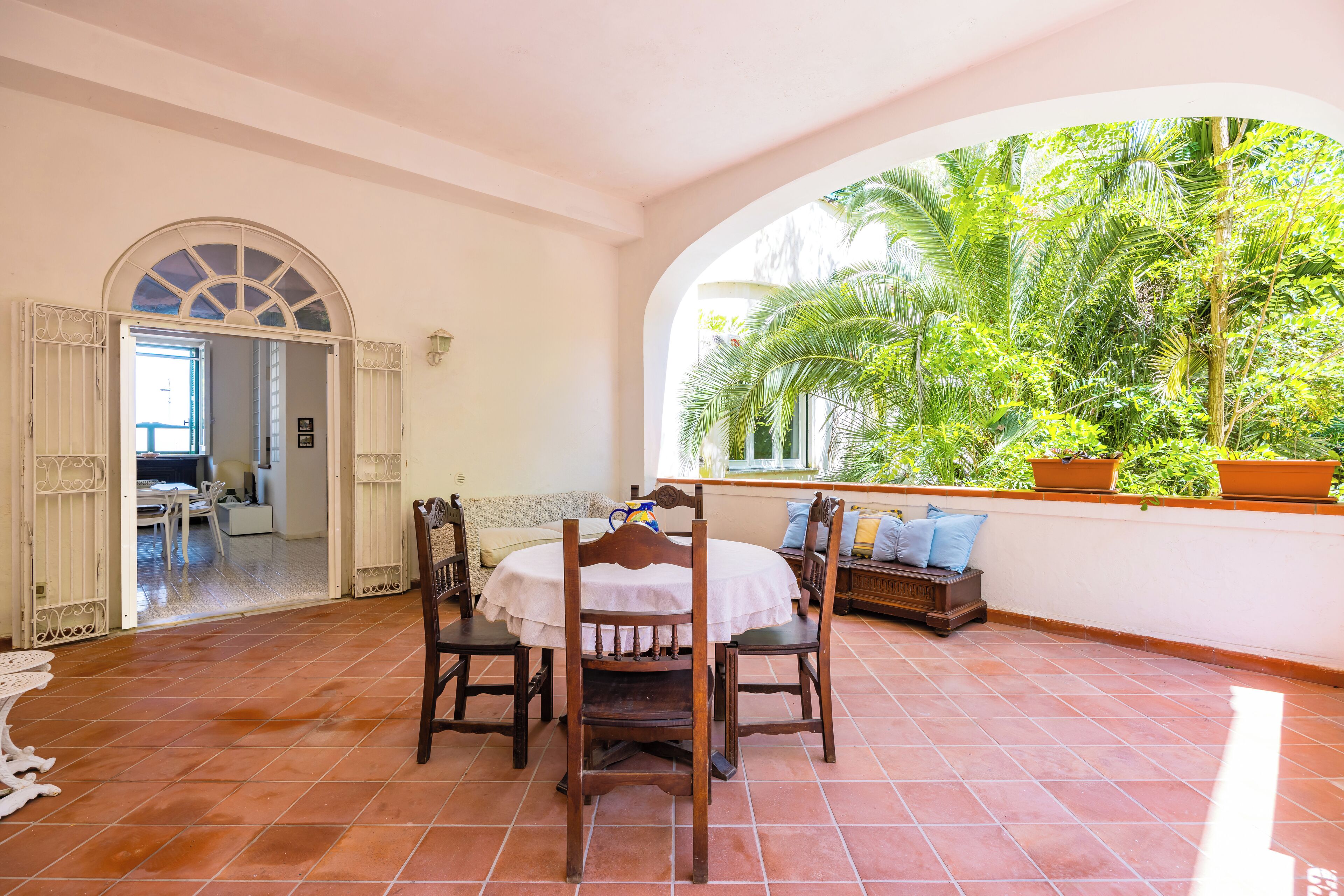 A dining area with a round table and chairs, a view of lush greenery through an arched window, and a tiled floor.