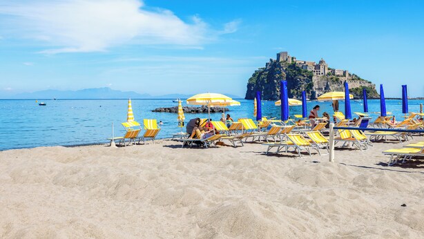 Une plage avec des transats et des parasols, un ciel bleu dégagé et, au loin, sur une colline, un château.