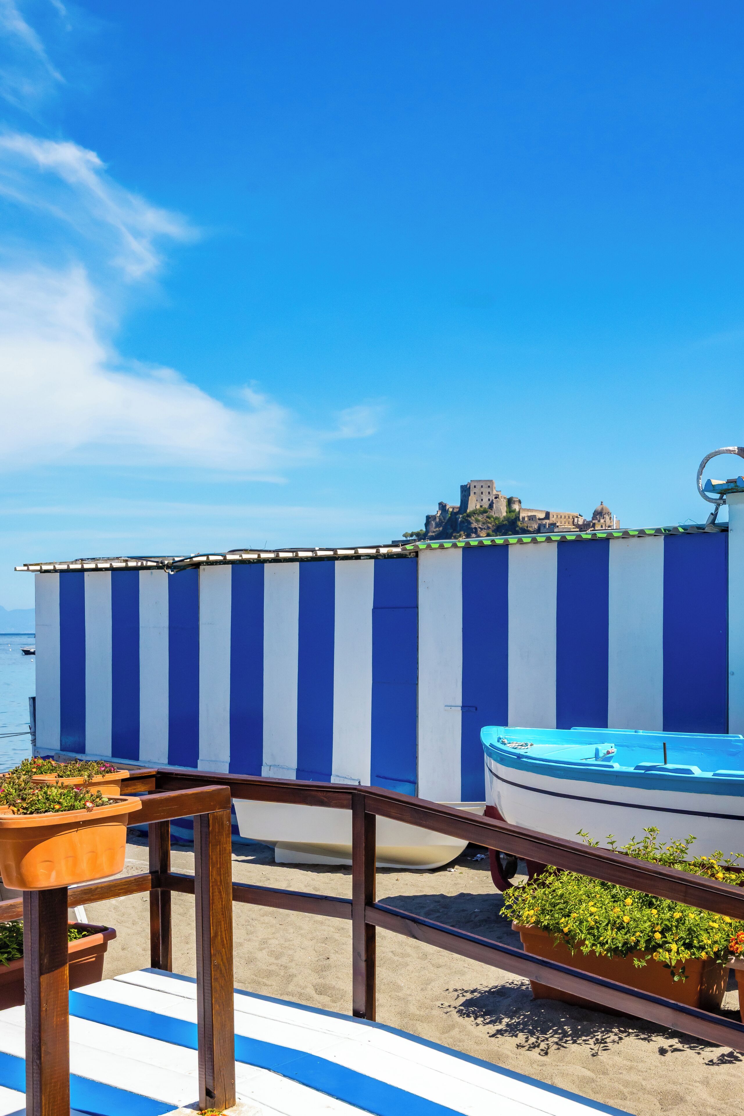 Eine Strandterrasse mit einer blauen und weißen Streifenwand, einem Boot und Blick auf eine Burg auf einem Hügel.