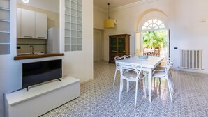 Un comedor con una mesa y sillas blancas, un televisor sobre un mueble blanco, y una ventana con vistas a palmeras.