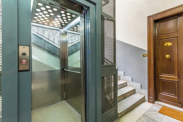 Un ascensor con puerta metálica y panel de control, una escalera con escalones de mármol, y una puerta de madera con pomo de latón.