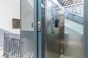 Interior de elevador com painel de comando, paredes metálicas e porta de malha.