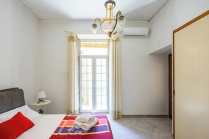Quarto de hotel com cama, mesa de cabeceira, luminária, lustre e janela com cortinas.