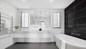 A modern bathroom with a freestanding bathtub, a double vanity with two sinks, and a large mirror above the vanity. - Walk to Waves - Bronte Stay (Bronte)