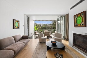 A modern living room with a sofa, a coffee table, and a fireplace. There is a balcony with outdoor furniture and a view of greenery.
