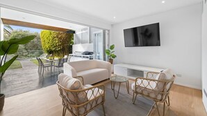A modern living room with a sofa, two wicker chairs, a coffee table, and a flat-screen TV mounted on the wall. - Walk to Waves - Bronte Stay (Bronte)