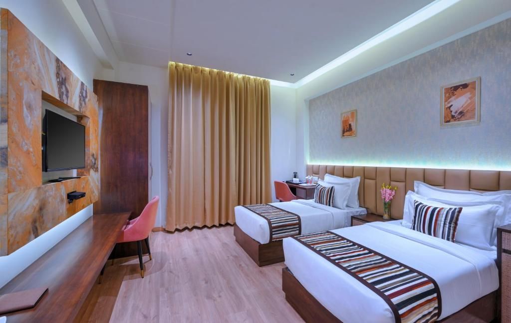 A hotel room with a flat-screen TV, two beds with striped bedding, a desk with a chair, and a large window with curtains.