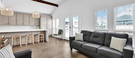 A spacious living area with a grey sofa, wooden flooring, and a kitchen with white cabinets and pendant lights.
