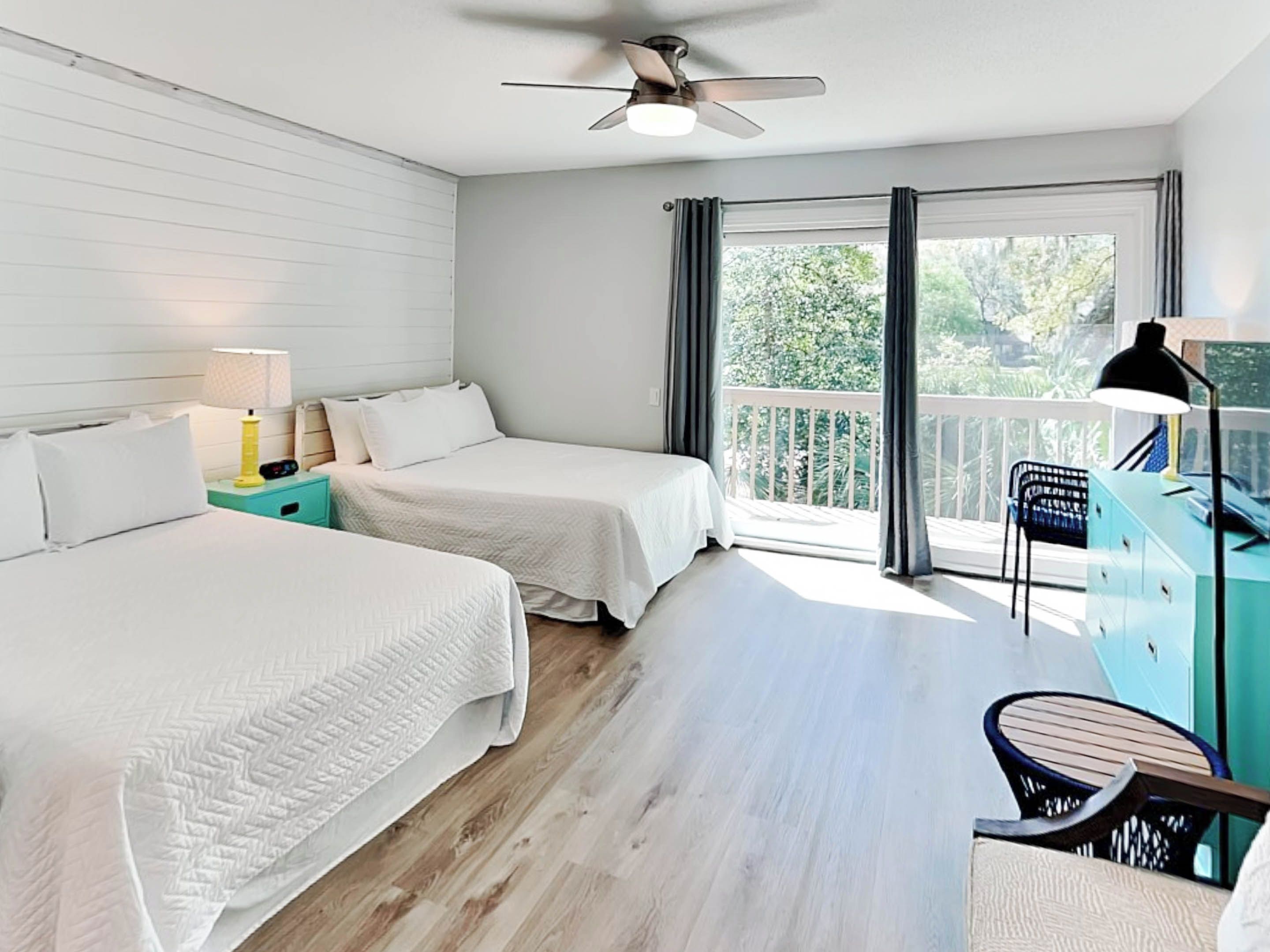 A hotel room with two beds, a desk, a chair, a ceiling fan, and a balcony with a view of greenery.