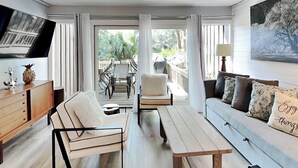 A modern living room with a sofa, a dining table, and a TV. There is a balcony with a table and chairs. - Water Oak Villas #2 by Hilton Head Retreats (Hilton Head Island)
