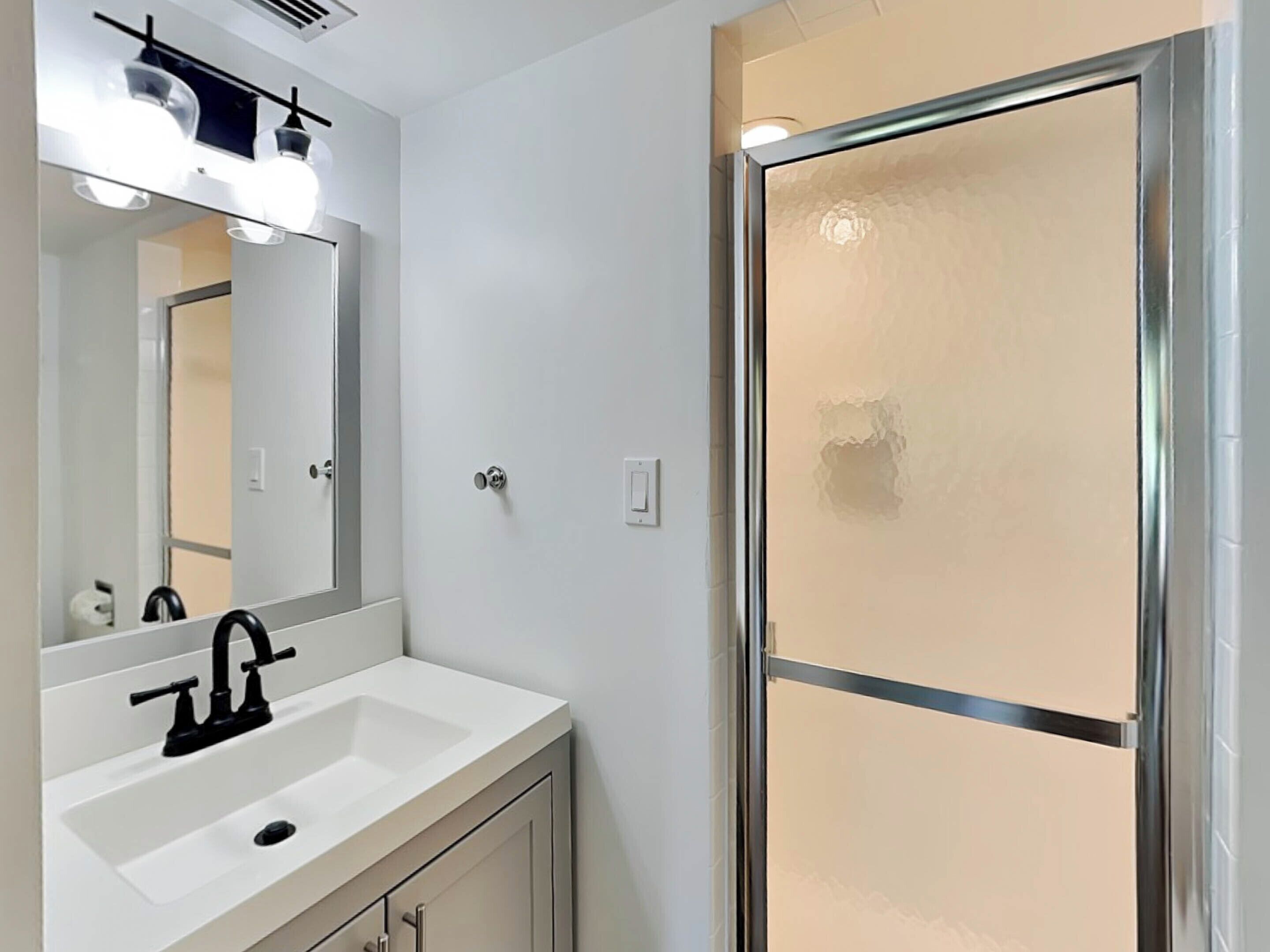 A bathroom with a white sink, black faucet, and a mirror above it. There is a shower with a glass door and a towel rack above it.
