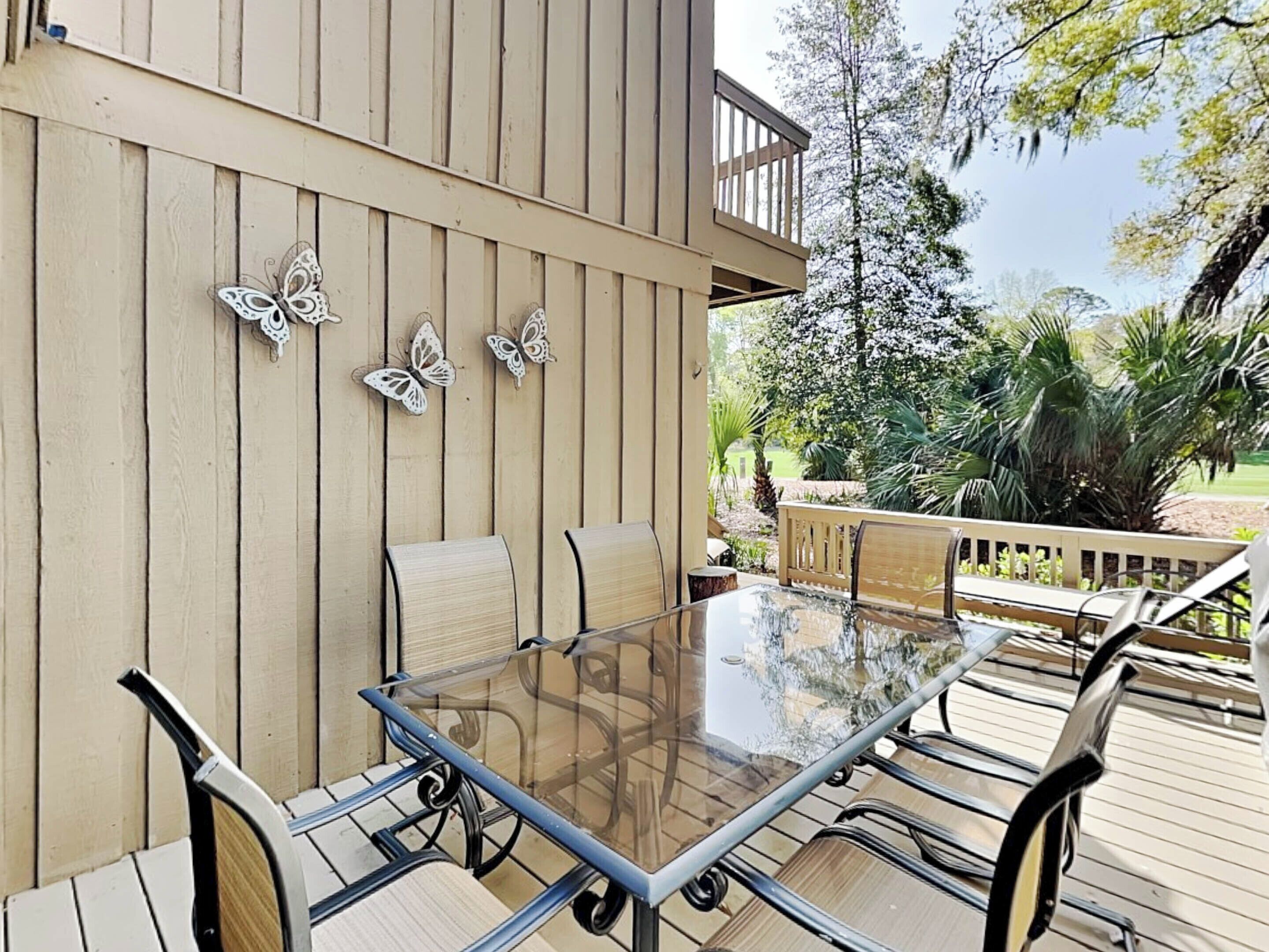 Eine Terrasse im Freien mit einem Tisch mit Glasplatte, umgeben von Stühlen, einem Holzboden und dekorativer Wandkunst mit Schmetterlingsmotiven.