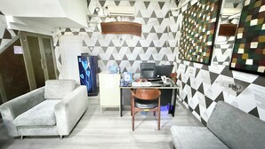 Una habitación pequeña con sofá, escritorio y silla. Hay un refrigerador, una computadora y una pared con diseños geométricos.