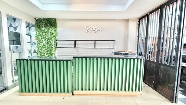 Un mostrador de recepción con una decoración verde y blanca, una placa con el nombre "KOTJIL" y un panel de vidrio con diseños geométricos.